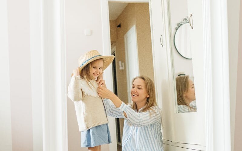 A mother assists her young daughter in trying on a hat while packing for a vacation indoors.