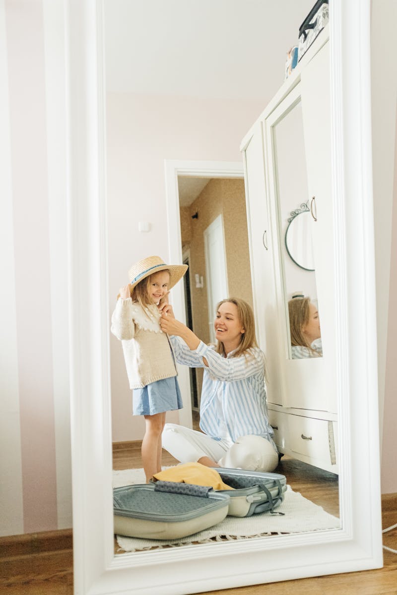 A mother assists her young daughter in trying on a hat while packing for a vacation indoors.